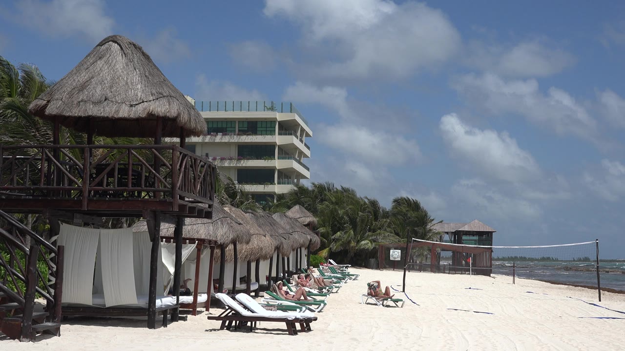 At 0:01, the Playa Del Carmen beach view holds on the raised palapa hut, rows of lounge chairs, a volleyball net near the water, and small beachgoers in the distance.