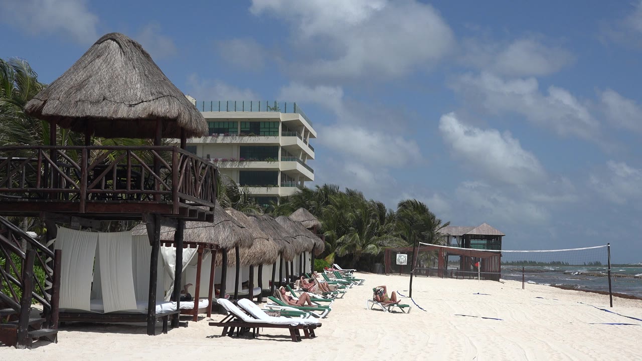 Thatched beach cabanas, loungers, people, ocean, and cloudy blue sky.