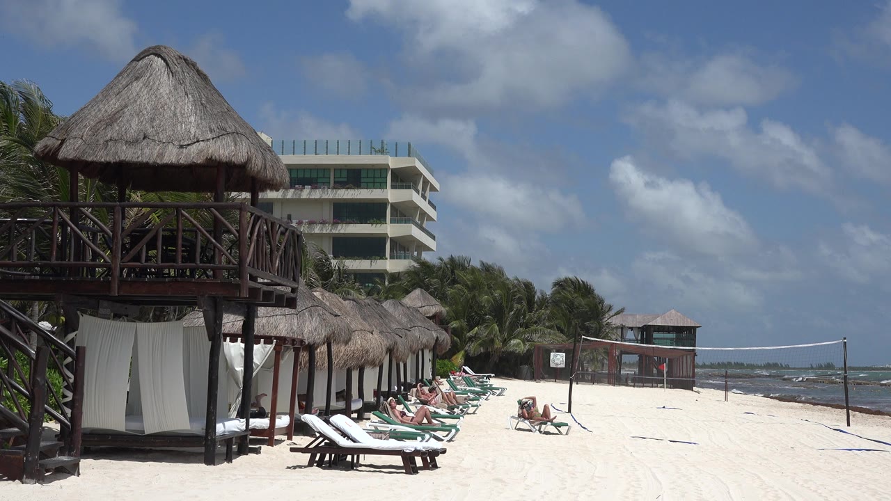 Playa del Carmen shoreline view with the thatched cabana still prominent, sun loungers occupied in the midground, and small waves visible beyond the volleyball net.