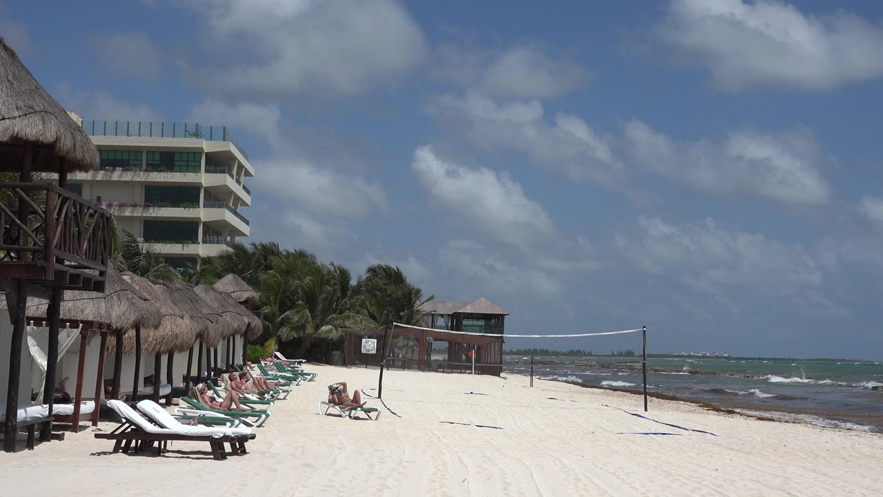 Opening frame of a Playa del Carmen beach with lounge chairs and thatched shade structures at left, people reclining near the sand, a volleyball net in the middle, and ocean water on the right.