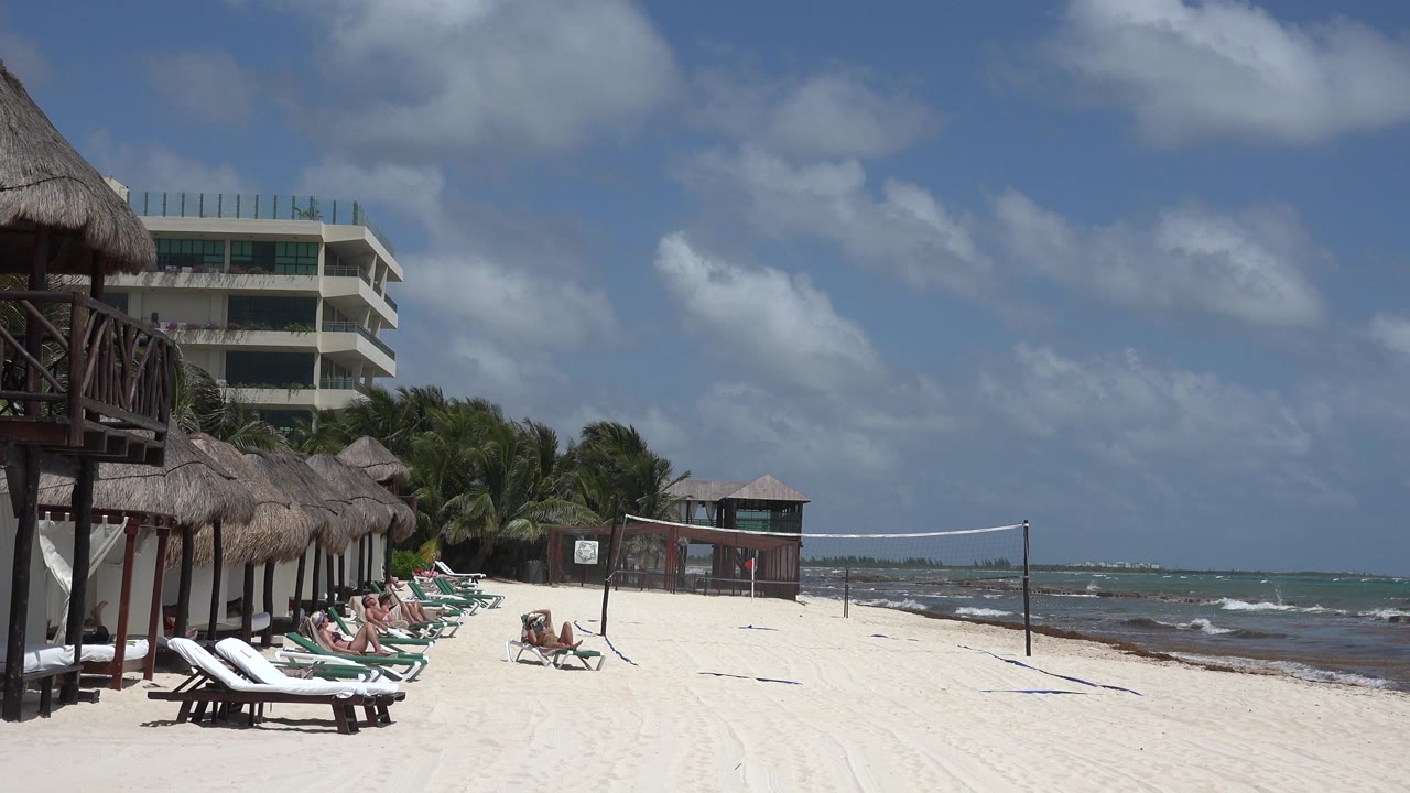 At 0:01, the Playa del Carmen shoreline remains framed with empty and occupied loungers on the left, the volleyball net across the sand, and small waves visible beyond the net.