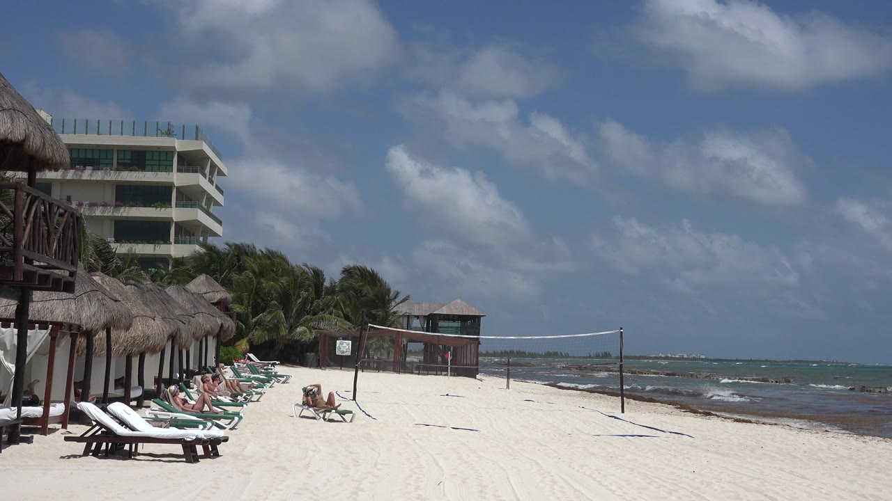 Sunny beach with reclining people, shaded loungers, volleyball net, and ocean horizon.