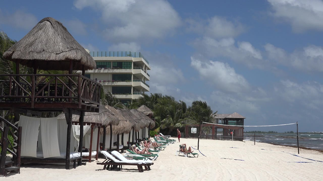 Beach loungers beside palapa shade structures and a volleyball net by the water.