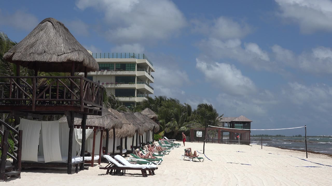 Playa del Carmen beach view one second in, showing the same palapa deck and resort-side building with people seated or standing near lounge chairs and the shoreline.