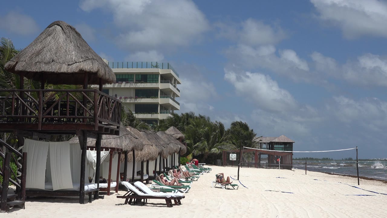 Playa del Carmen beach view with the thatched structure still dominant on the left, a row of lounge chairs below it, cabanas along the sand, rough surf in the distance, and no identifiable people.