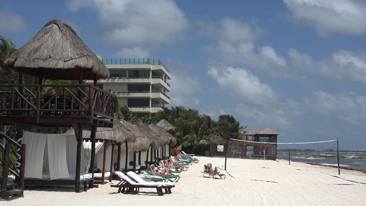 Beach loungers, sunbathers, thatched hut, sea, and volleyball net.