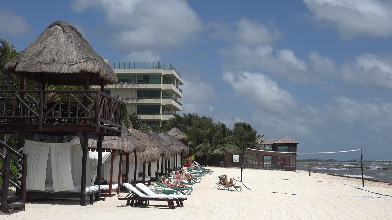 Playa del Carmen beach frame with the dark wooden hut structure still dominant at left, a row of loungers with visible people at center, and wind-textured water beyond the volleyball net.
