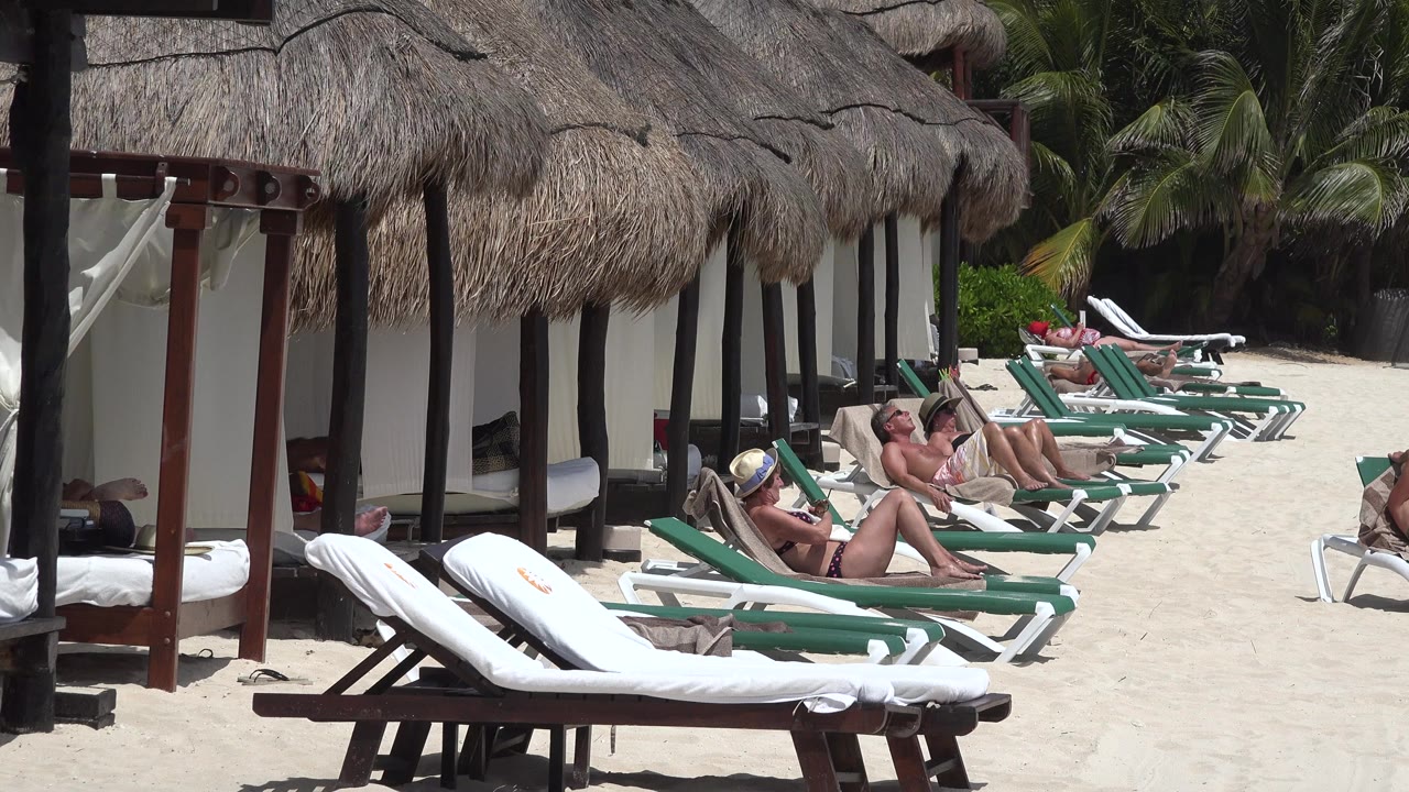 At Playa del Carmen, adults recline on green-and-white beach loungers beside thatched shade structures on bright white sand.