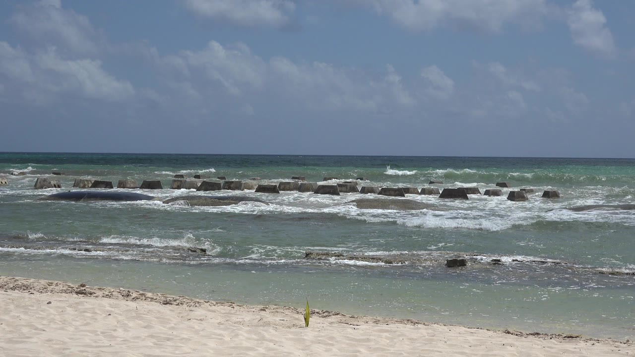 Opening frame of the Playa del Carmen shoreline with pale sand in front, green-blue water, breaking waves on dark blocks, a level horizon, and no visible people.