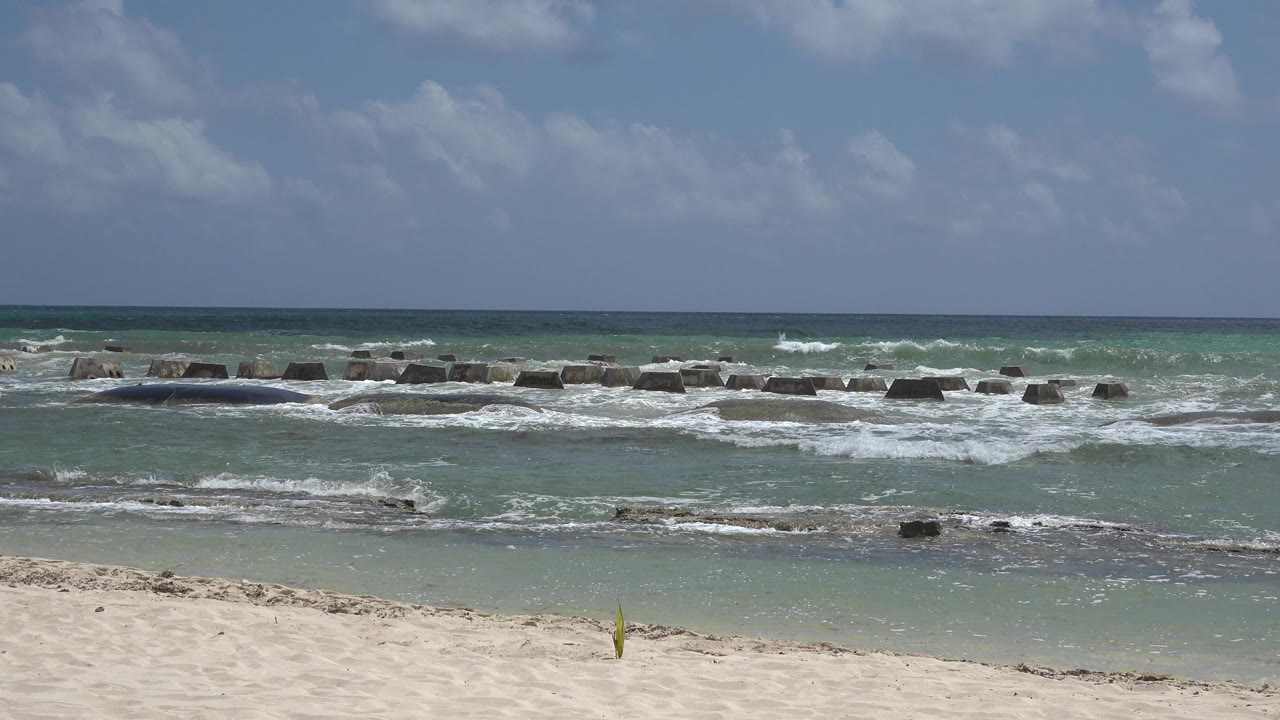 Playa del Carmen surf spreads across shallow water as white foam gathers near the dark rock line, with sand at the bottom of frame and no visible people.