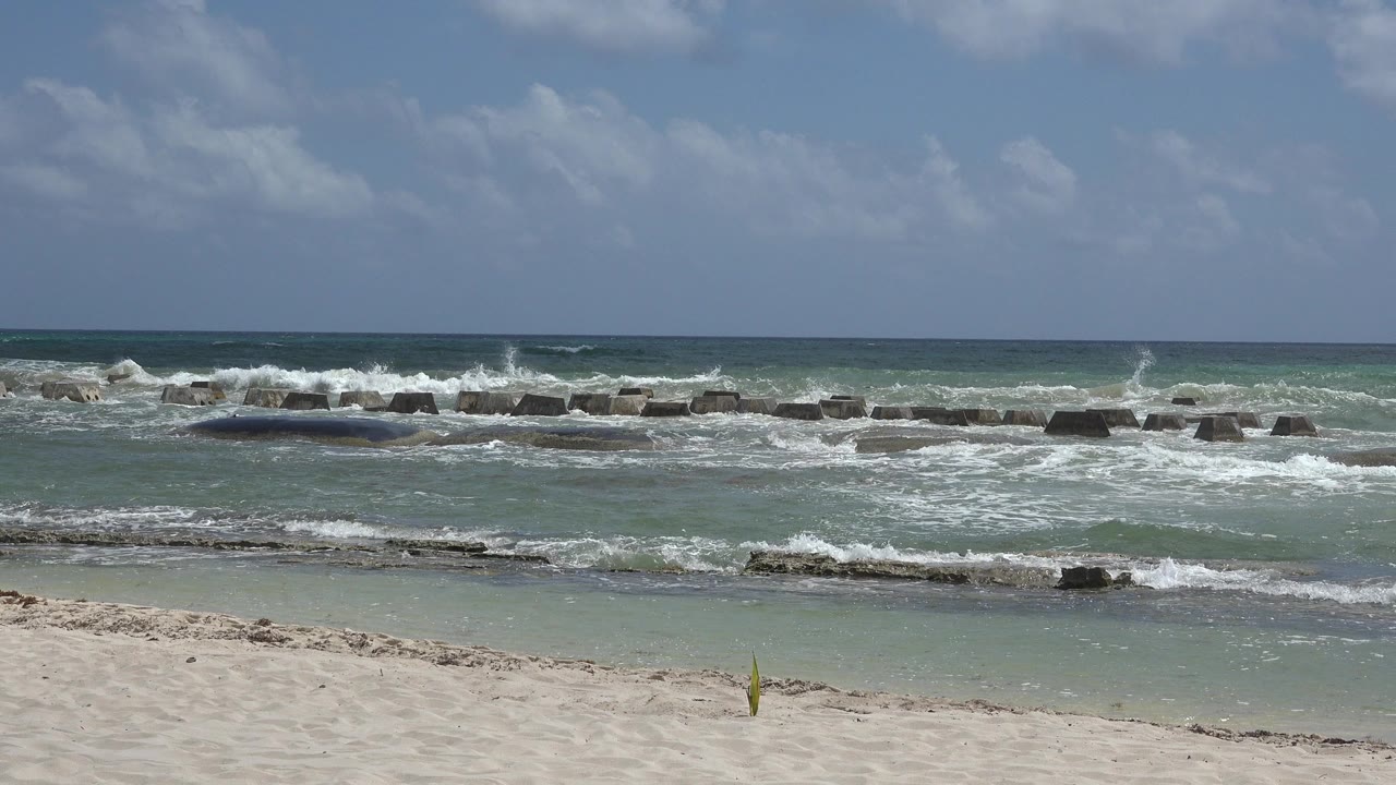Playa del Carmen shoreline with pale sand in front, green-blue water, white surf hitting low breakwater rocks, scattered clouds, and no visible people.