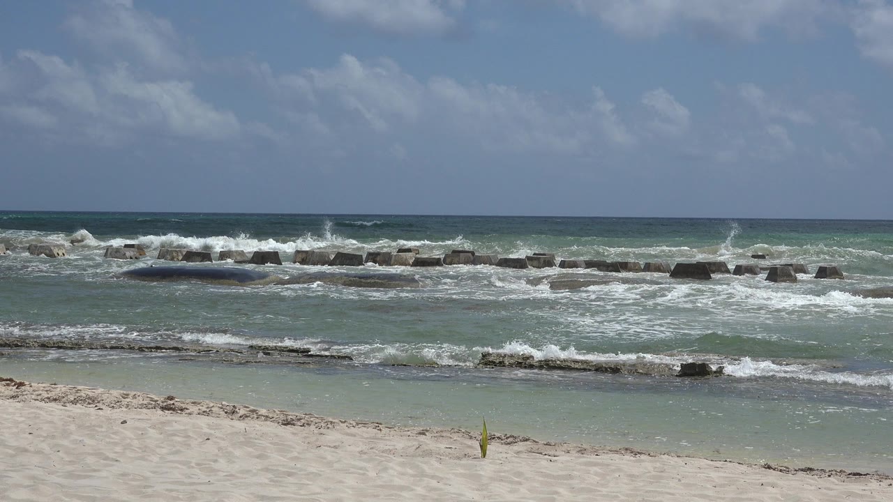 Playa del Carmen beach view with foam spreading across shallow water, low dark barriers across the midground, open horizon, cloud patches, and no visible people.
