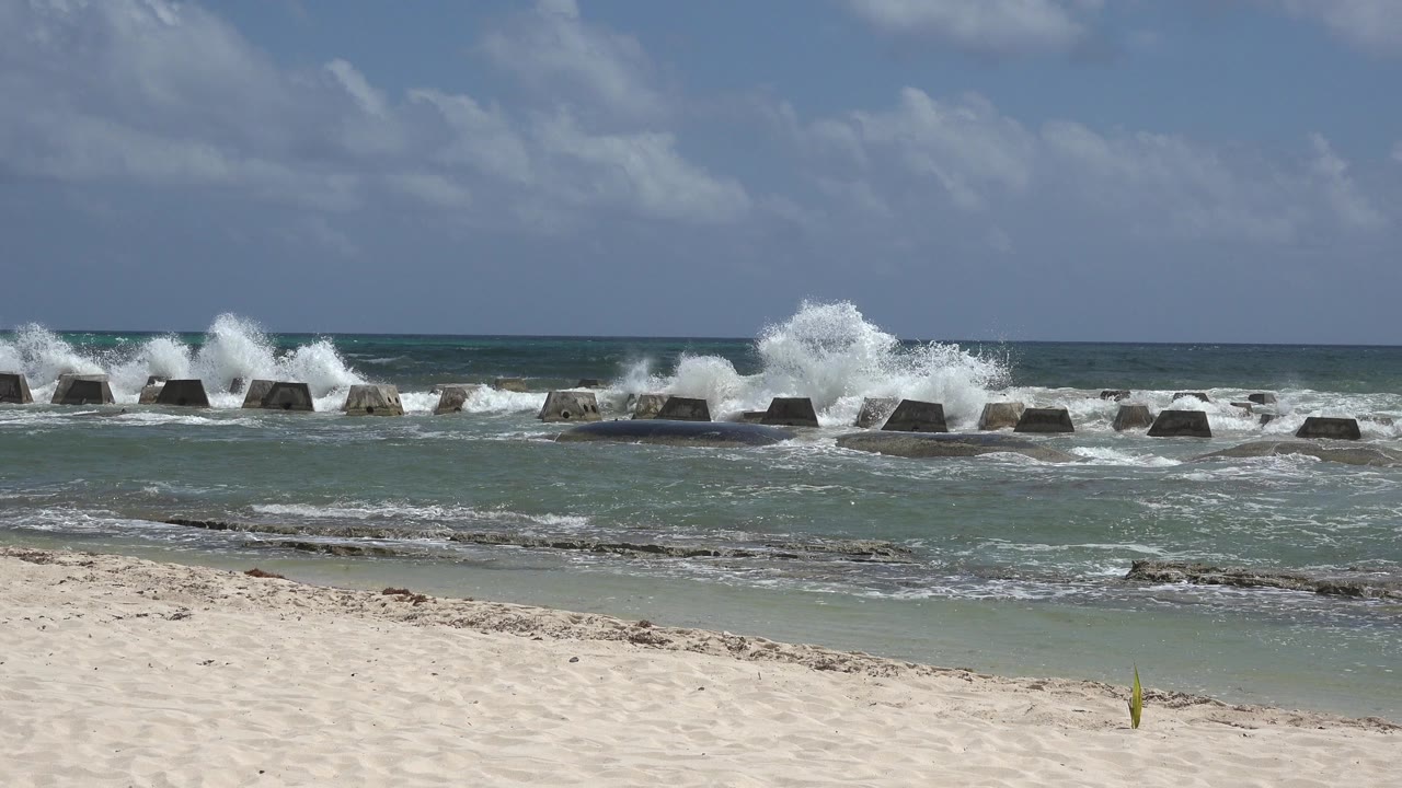 At the opening moment in Playa del Carmen, Mexico, pale sand leads to green-blue surf as a wave splashes over concrete breakwater blocks, with no visible people.