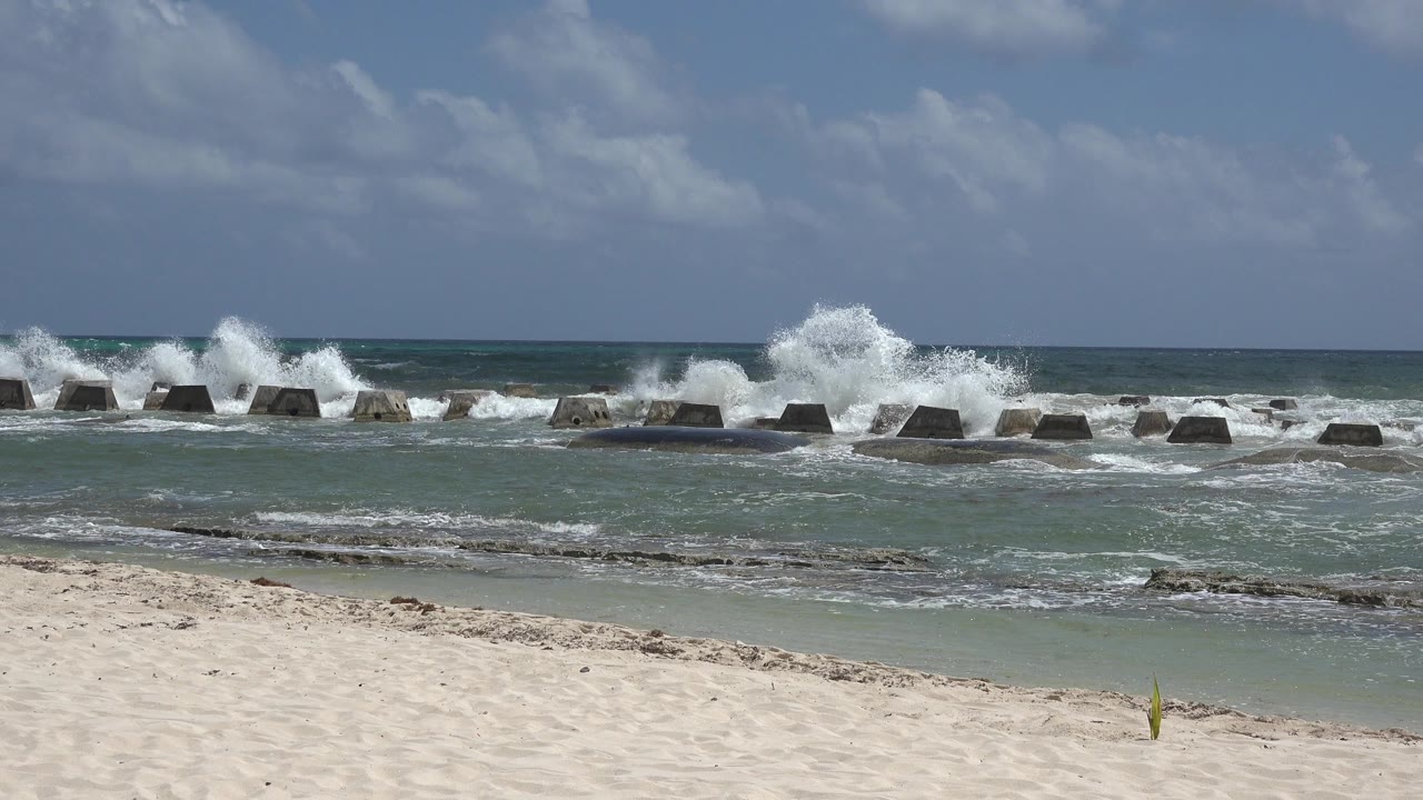 One second into the Playa del Carmen clip, a taller burst of white surf hits the offshore concrete barriers behind the shallow water, with no visible people.