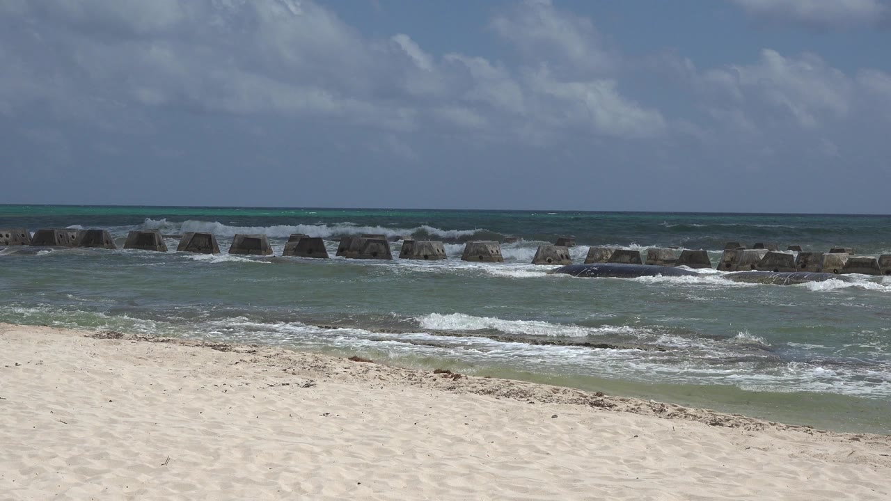 Sandy beach with surf and concrete breakwater blocks offshore.