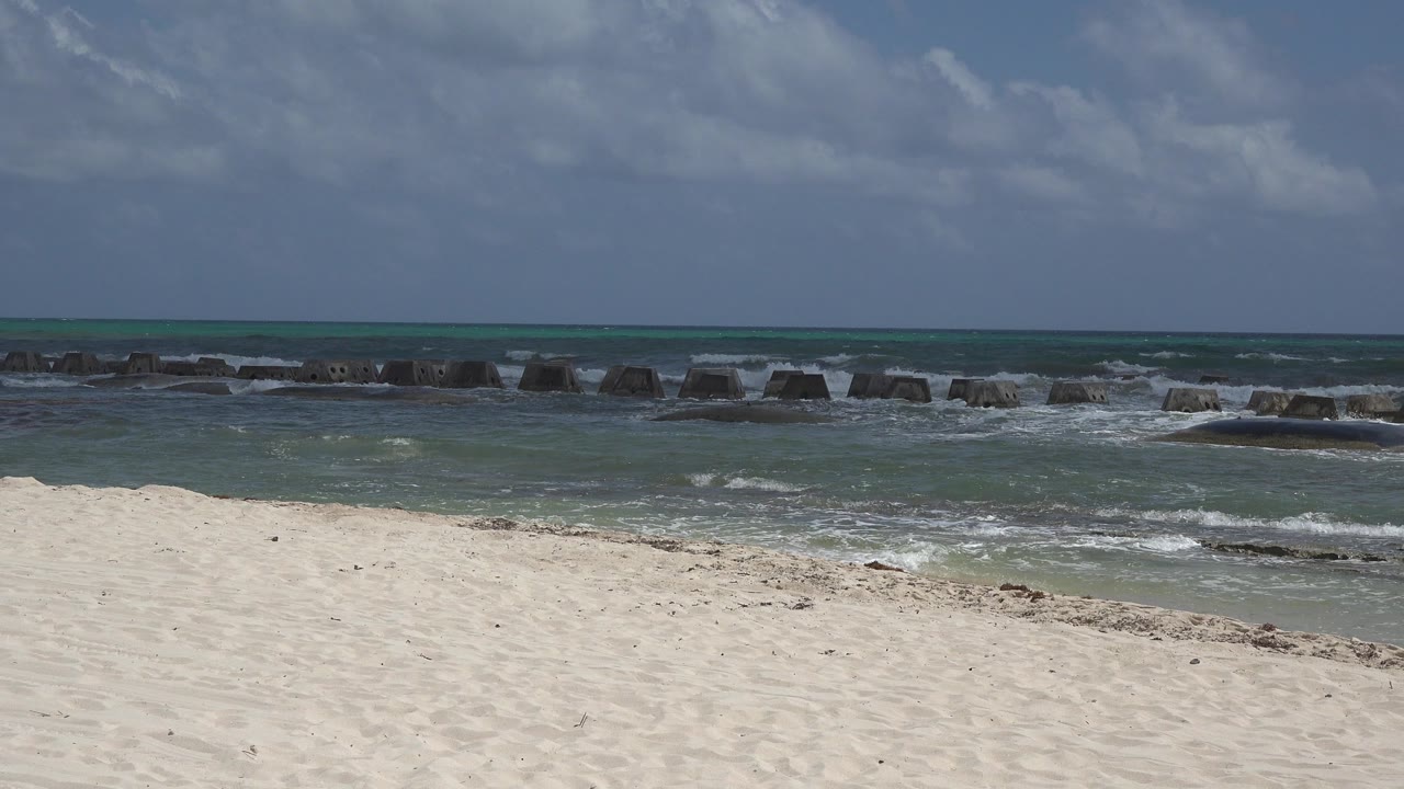 Opening frame of a Playa del Carmen beach with pale sand in front, small waves breaking around dark offshore rocks, cloudy blue sky, and no visible people.