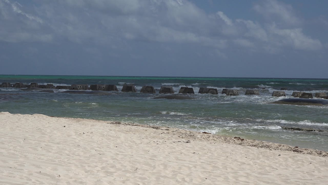 Playa del Carmen shoreline at 0:01 with a wide sand foreground, foamy waves reaching the rocky midground, a level horizon, and no visible people.