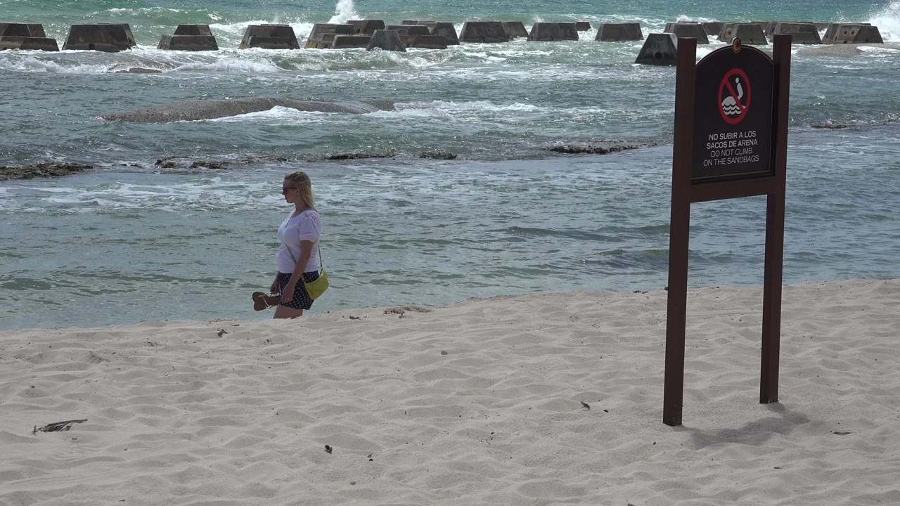 A blond girl stands on the sand in Playa del Carmen, Mexico, with rough turquoise surf behind her and a Spanish warning sign near the right side of the frame.