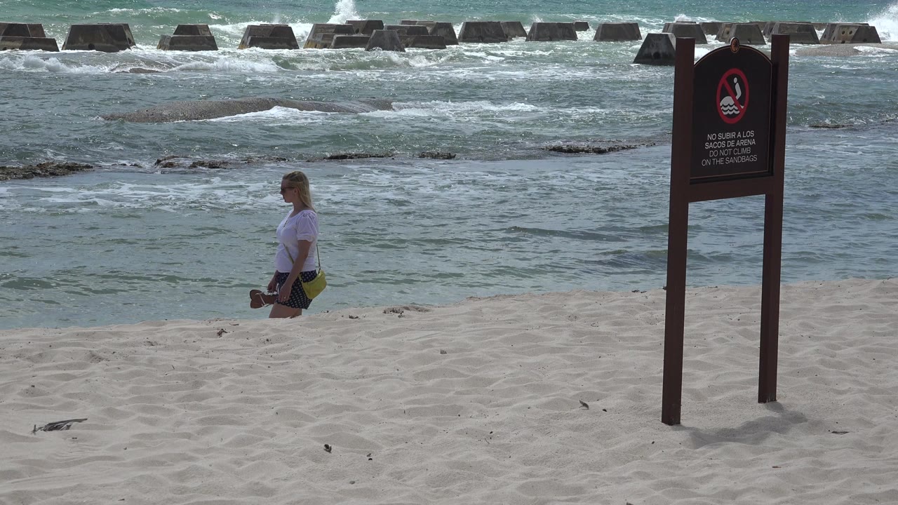 The girl remains on the sandy beach in Playa del Carmen, Mexico, while whitewater spreads across the water and the warning sign stands in the right foreground.