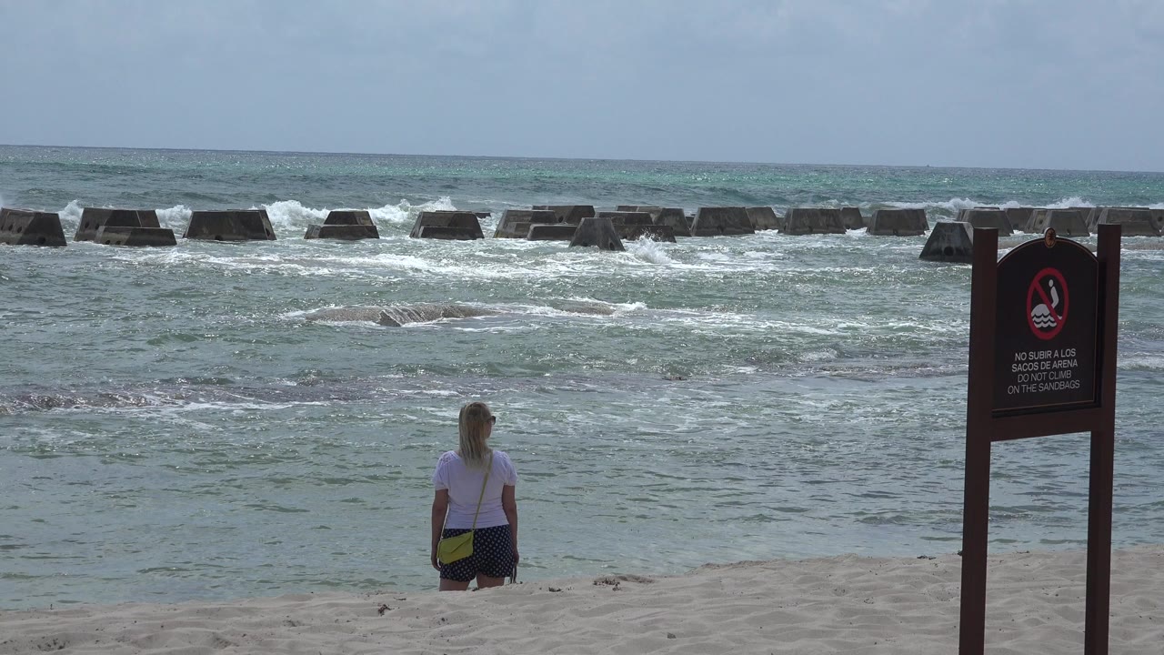 At 0:01 in Playa del Carmen, Mexico, the woman remains near the lower center facing the water while white surf breaks against concrete barriers offshore.