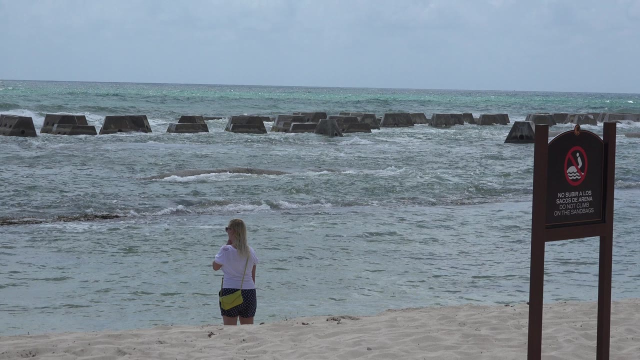 Woman standing on sandy beach near waves and a warning sign.