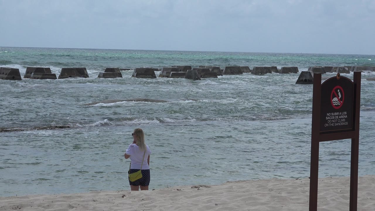 The woman remains near the waterline at Playa del Carmen, Mexico, while small white waves cross the midground and the dark beach warning sign stands at the right edge.