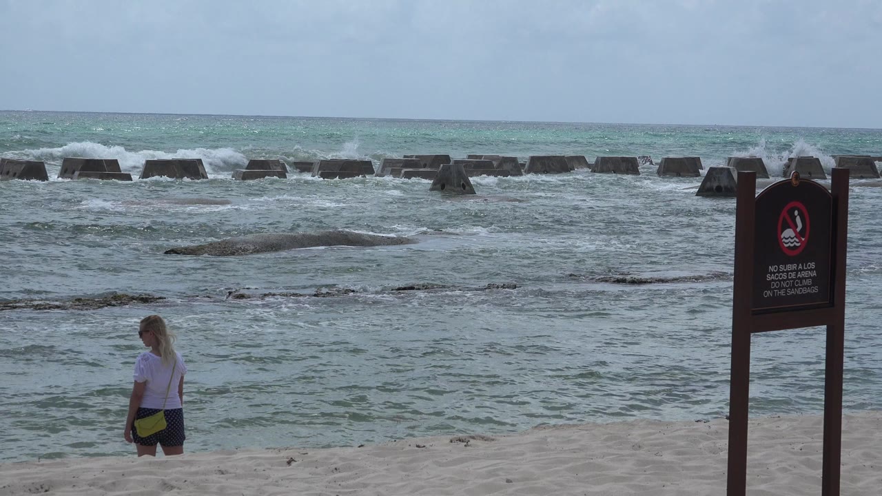 In Playa del Carmen, Mexico, a blonde woman stands near the waterline while waves roll behind a dark beach warning sign at the start of the clip.