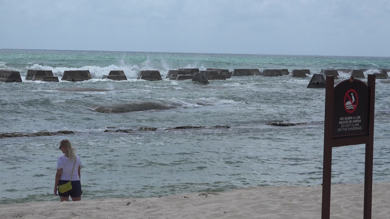 Beach warning sign beside a woman looking toward choppy surf.