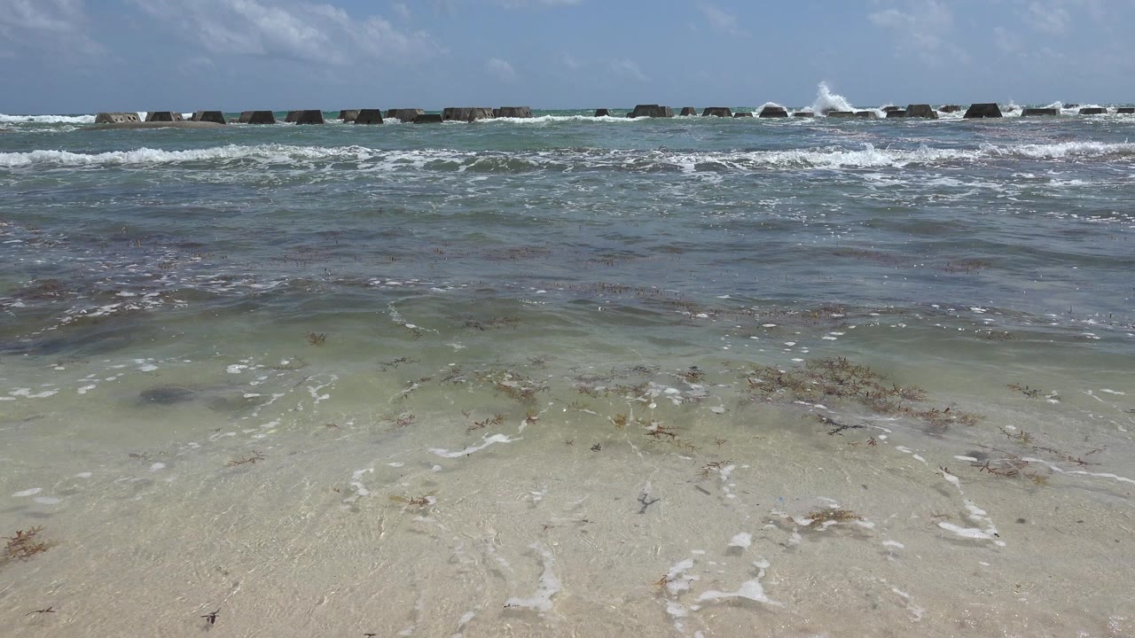 At 0:00 in Playa del Carmen, Mexico, shallow water spreads across pale beach sand with a dark rock breakwater near the horizon and no visible people.