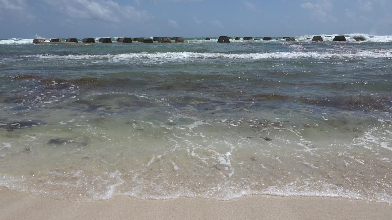 At 0:00 in Playa del Carmen, shallow surf rolls over pale sand beneath a blue sky, with dark rock barriers across the horizon and no visible people.