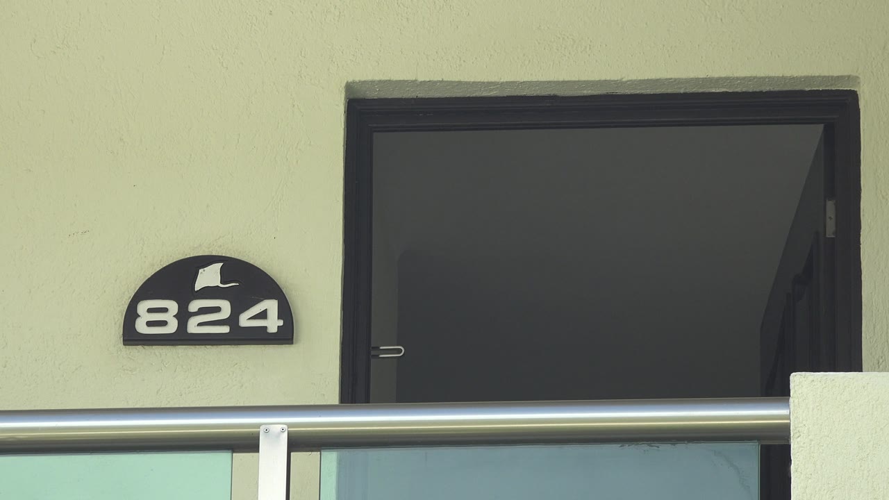 Close framing on the Playa del Carmen apartment facade shows the 824 sign left of a dark open doorway, a horizontal balcony rail, and no visible people.