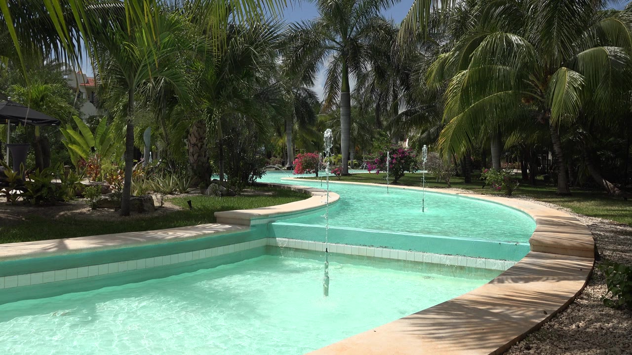 Opening view of a turquoise decorative pool curving through tropical landscaping in Playa del Carmen, Mexico, with palms, water jets, a tan walkway, and no visible people.