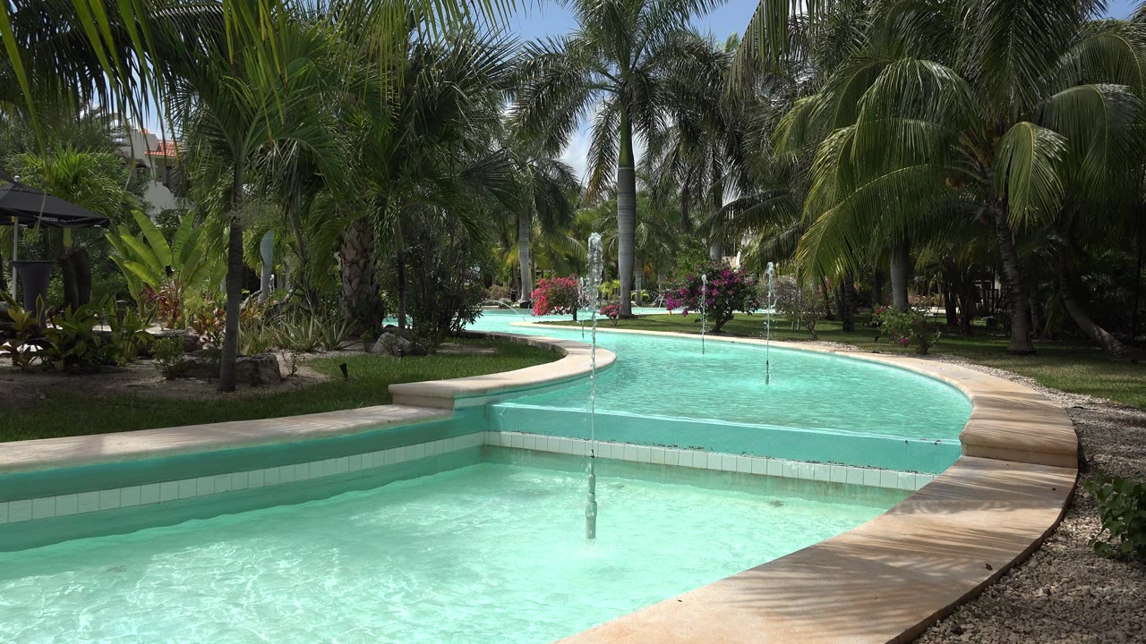 Opening view of a circular fountain in Playa del Carmen, Mexico, with pale turquoise water, a curved walkway, dense palms, and no recognizable people visible.