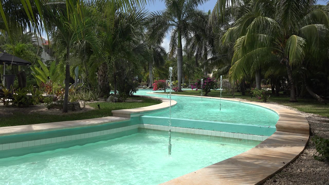 Opening frame of a turquoise infinity pool in Playa del Carmen, Mexico, with a curved beige edge, palms, lawn, water jets, and no visible people.