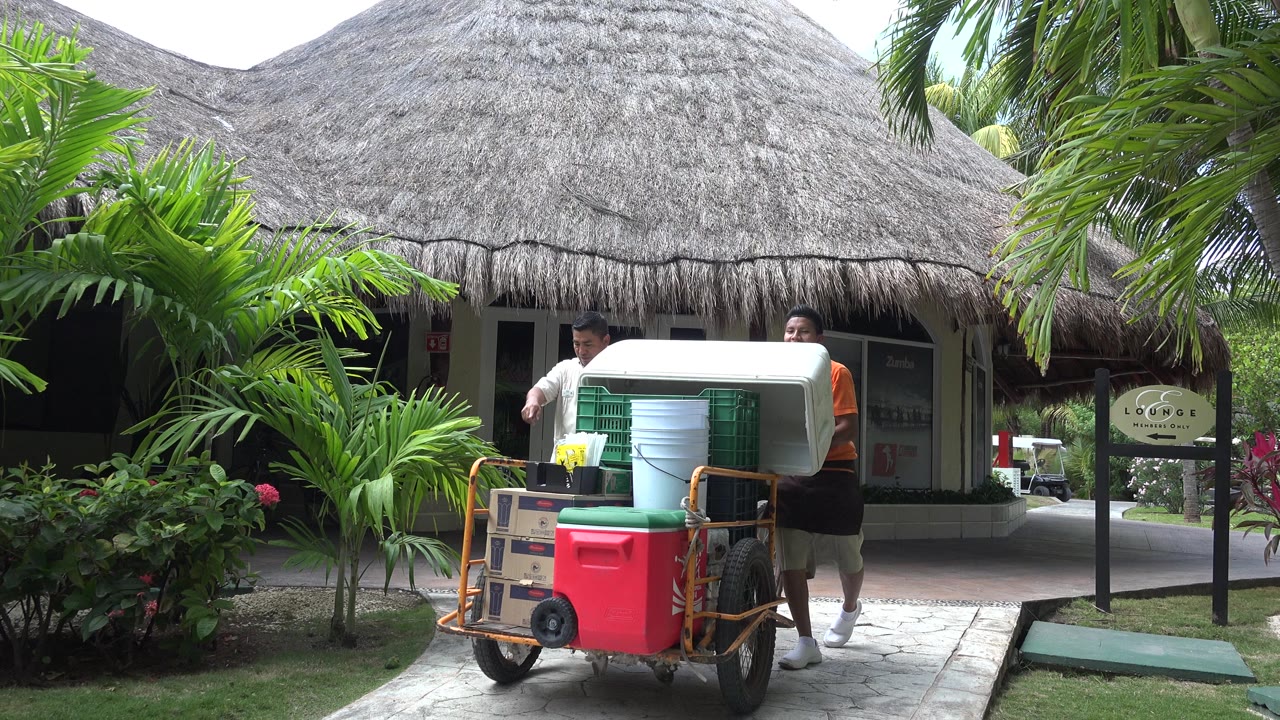 Two men push a utility cart loaded with coolers and crates past tropical plants and a thatched hut in Playa Del Carmen, Mexico.