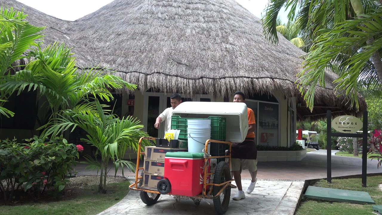 The loaded cart with coolers, crates, cups, and buckets fills the foreground while two workers stand behind it at the resort hut in Playa Del Carmen.