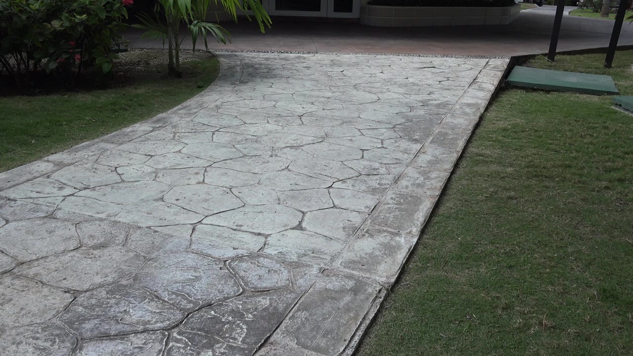 The pale stone pavement runs from the lower frame toward a shaded planted area in Playa Del Carmen, Mexico, with lawn on the right and no visible people.