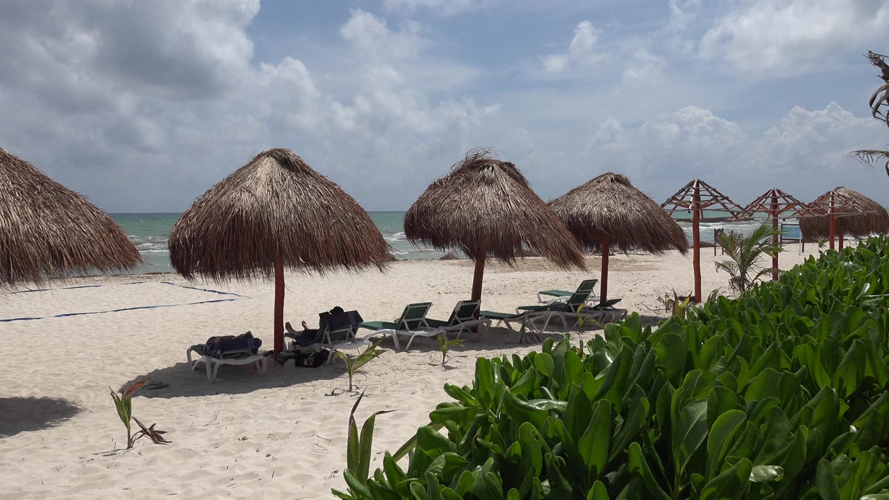 Opening frame of a Playa del Carmen beach with thatched parasols over empty loungers, green plants at right, pale sand, turquoise water, scattered clouds, and no visible people.
