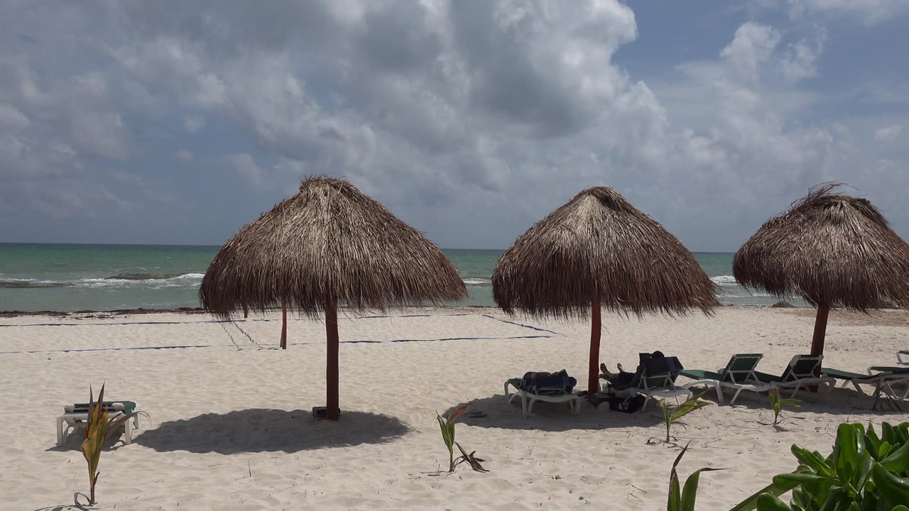 Opening frame of a Playa del Carmen beach with three thatched umbrellas, lounge chairs on pale sand, low greenery at the lower right, ocean in the background, and no visible people.