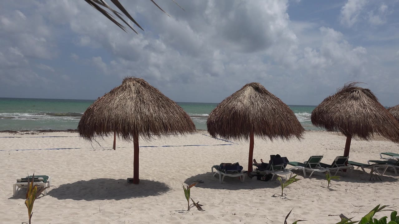 Playa del Carmen beach frame with three prominent thatched umbrellas, several lounge chairs, low surf behind them, and indistinct people reclining in the shaded seating area.
