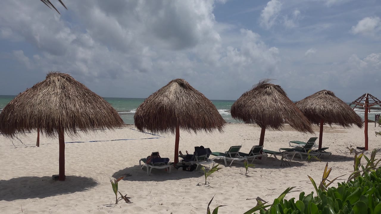 Beach palapas, lounge chairs, sand, and ocean under a cloudy sky.