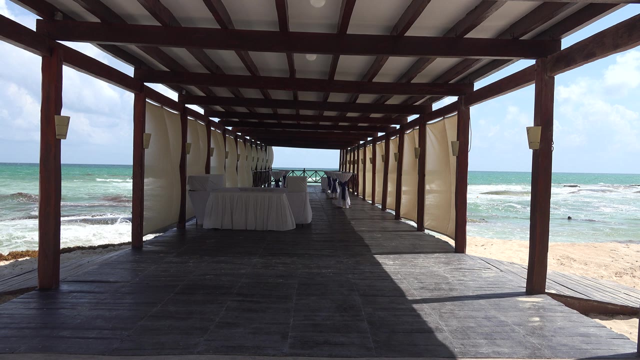 A shaded wooden walkway in Playa del Carmen faces white sand and breaking ocean waves, with banquet tables under the roof and no visible people.
