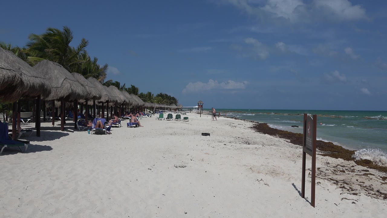Playa del Carmen beach at the start of the clip, with white sand in front, palapa umbrellas along the left, small groups of people in the distance, and low waves on the right.