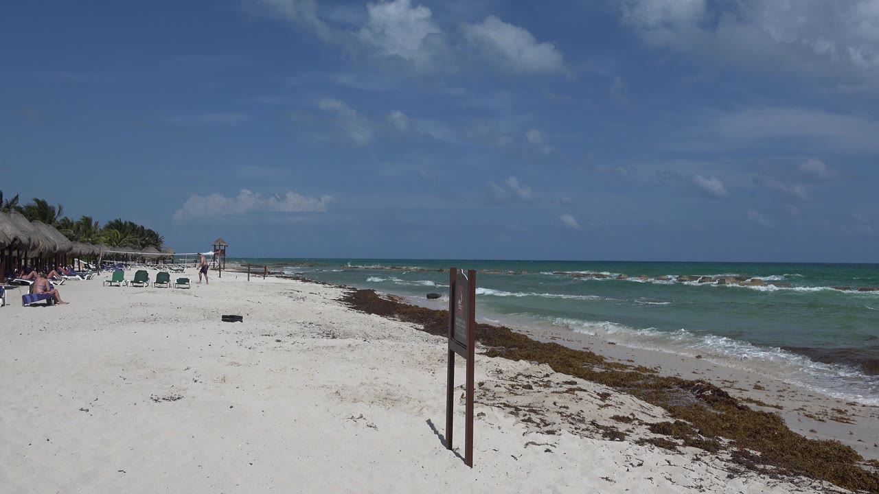 Waves roll beside a sandy beach with chairs and palapa shade in the distance.