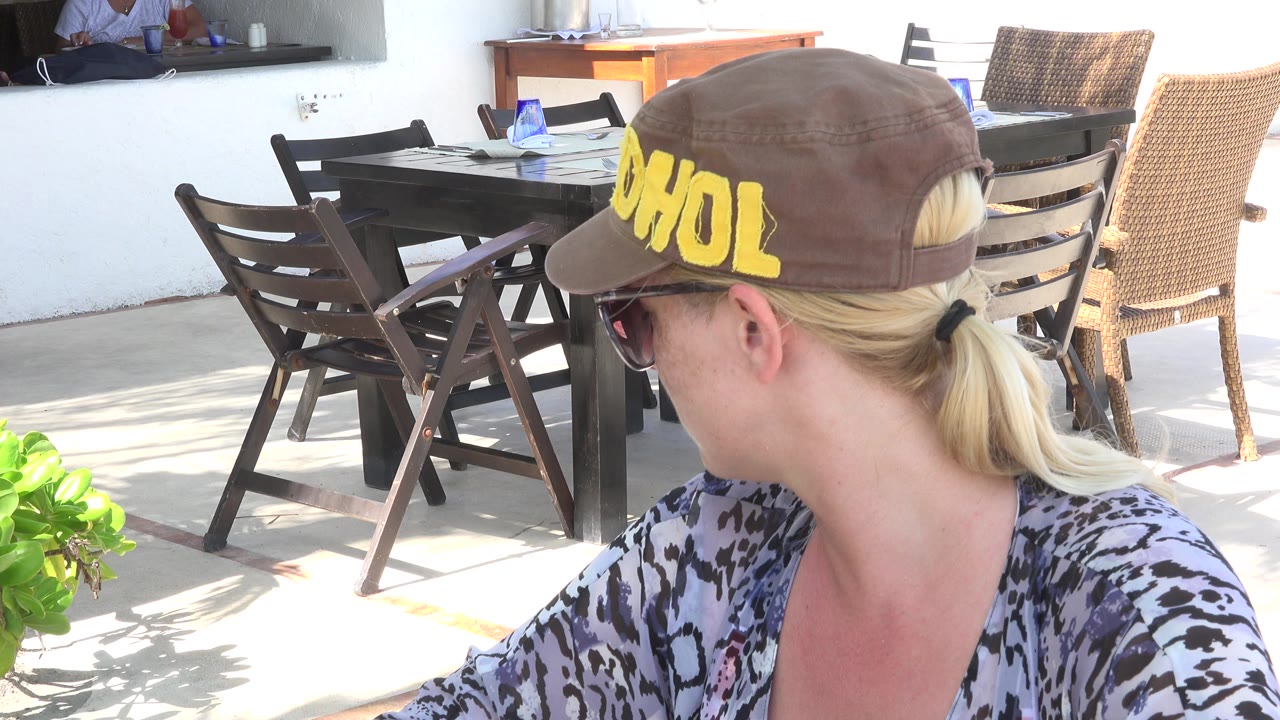 Blonde woman in sunglasses and cap looking over her shoulder at a patio table.