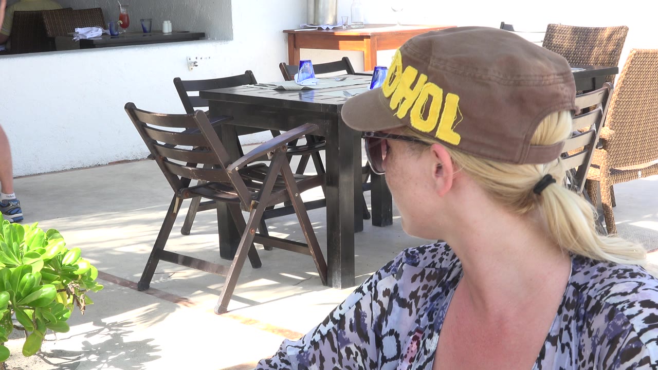 In Playa del Carmen, a blonde woman in a brown cap and sunglasses is seen from behind near a dark patio table, wooden chairs, and bright green plants.