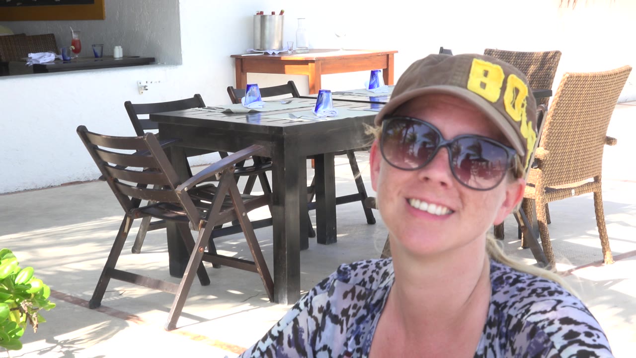 Smiling woman in sunglasses seated at an outdoor restaurant table.