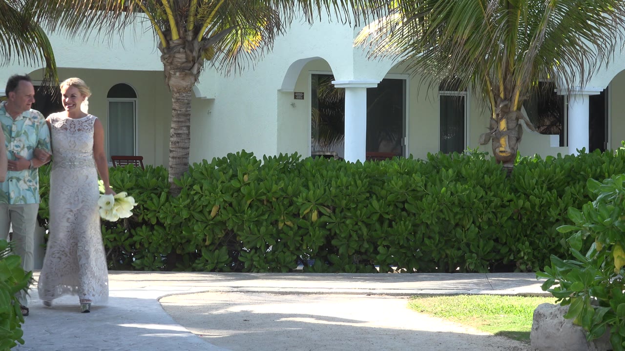 A newly married couple in Playa del Carmen walks from the left edge beside green hedges, palm trunks, and white resort arches in bright side light.