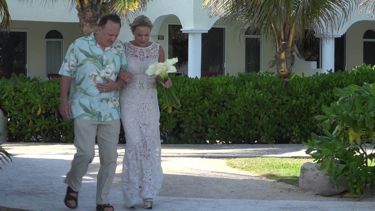 In Playa Del Carmen, Mexico, a bride in a white lace dress walks arm in arm with a man in a light floral shirt beside green hedges and palm trees.