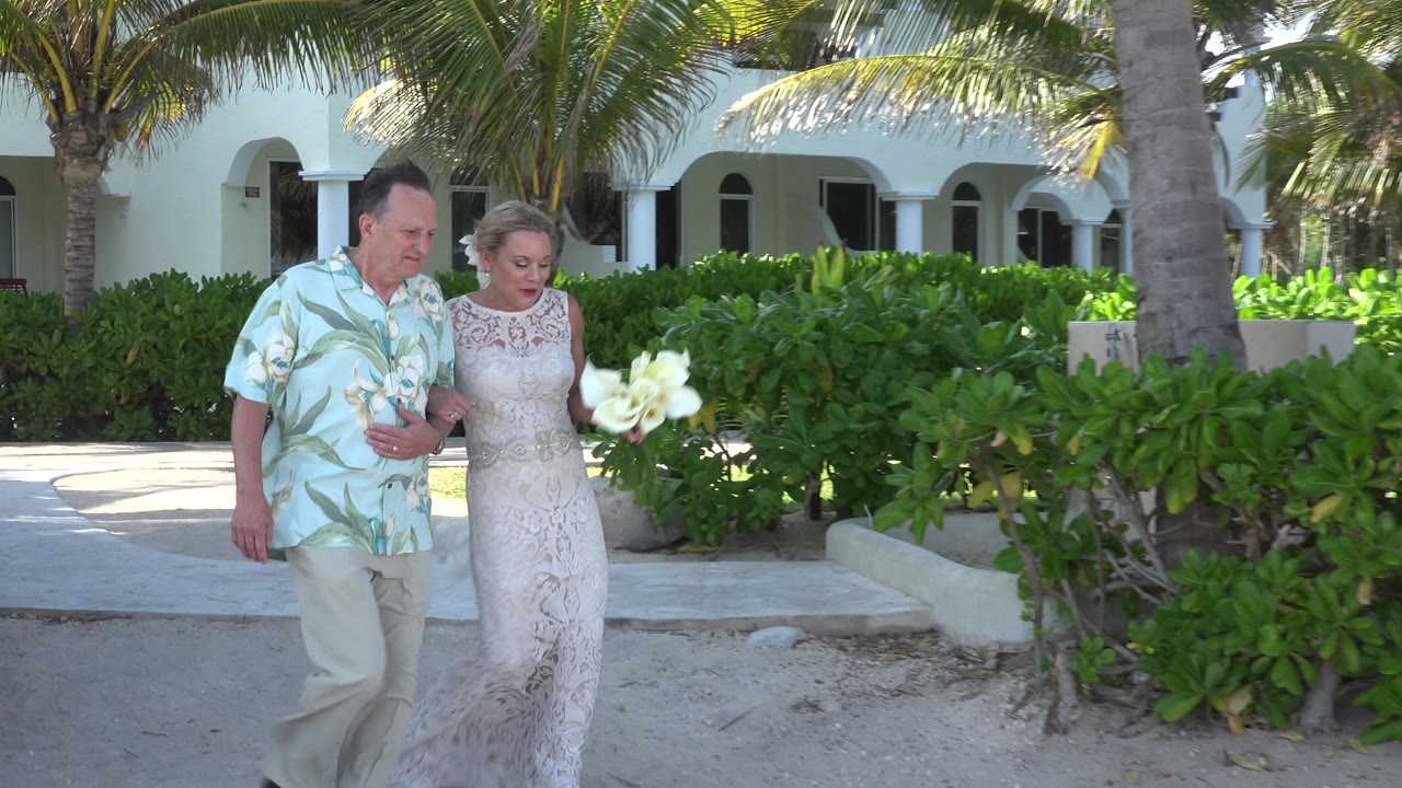 The newly married couple continues along the sandy path in Playa del Carmen, Mexico, with the woman's white flower bouquet held forward against green foliage.